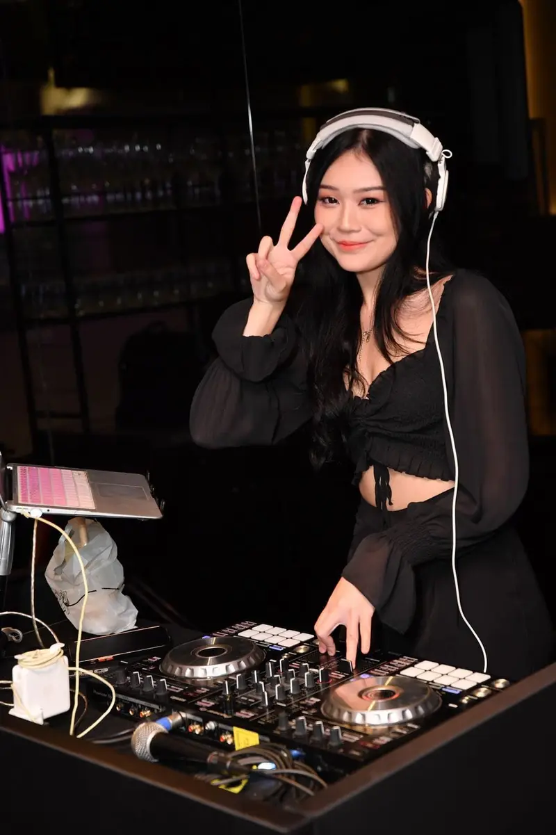 Female DJ in Singapore with white headphones smiling behind a DJ controller at an evening event
